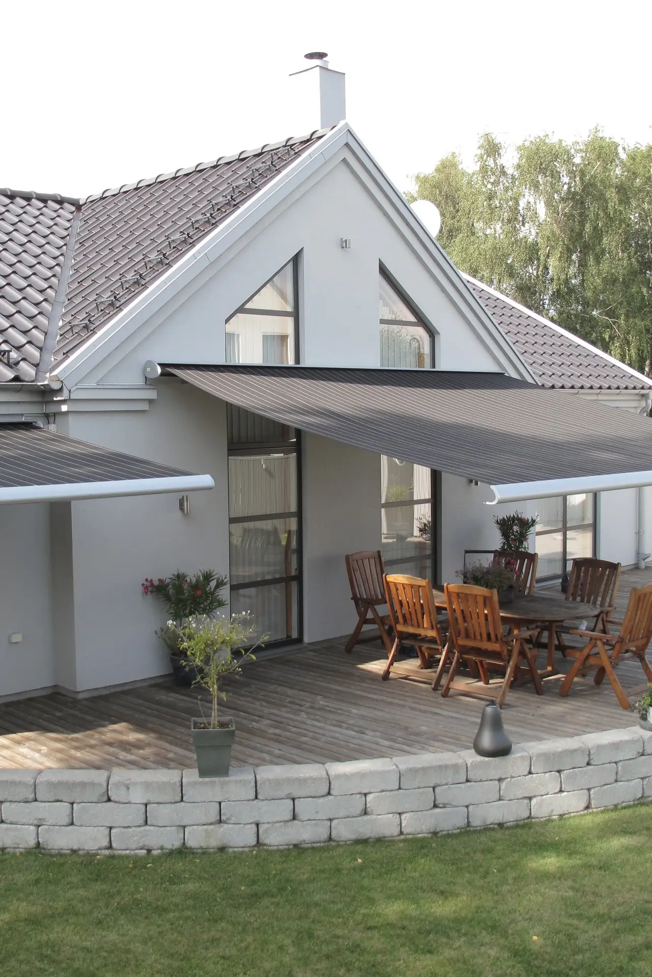 Terrassmarkis i grått över en altan med trädgårdsmöbler vid en vit villa med tegeltak